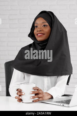 Portrait de belle femme musulmane portant le hijab noir et chemise blanche assis attable dans Office et de boire du café chaud. Jolie femme africaine à la recherche de côté, souriant et posant pendant les pauses. Banque D'Images