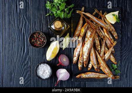 Close-up d'éperlans fumé à froid sur un plateau en ardoise noire avec du persil, des épices et des quartiers de citron, sur une table en bois, vue de dessus, flatlay, sp Banque D'Images