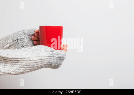 Woman's hands holding chandail confortable dans un rouge tasse de café, thé ou chocolat chaud contre le mur blanc. Banque D'Images