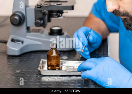 L'examen de l'assistant de laboratoire mâles dans des échantillons de biomatériaux d'un microscope. Jusqu'Cllose les mains dans des gants en caoutchouc bleu ajuster microscope. Banque D'Images