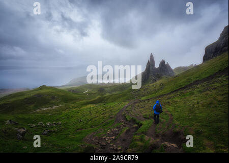 Old Man Storr. Le Storr sous un ciel nuageux dans un jour de pluie. Homme avec veste bleue de la randonnée à travers l'herbe sous la pluie. Île de Skie, en Écosse. Banque D'Images