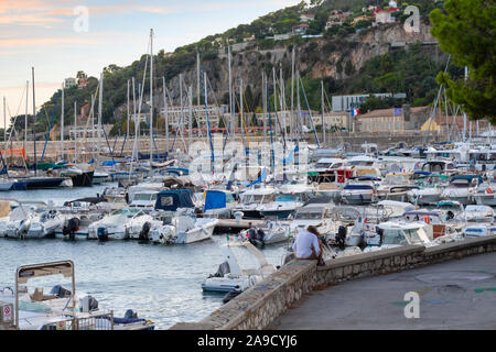 Une jeune personne aux cheveux longs est assis seul sur un mur de pierre le long de la marina au coucher du soleil à Villefranche-sur-Mer, France, sur la côte d'Azur. Banque D'Images