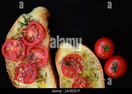 Deux tranches de pain blanc italien avec l'huile d'olive verte et de tranches de ​​Tomato avec l'origan à côté des tomates rouges sur un fond sombre Banque D'Images