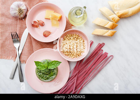 Rose brut fettuccine et sauce pesto avec des ingrédients : les noix de pin, le basilic, l'huile d'olive, le parmesan, l'ail rose en plaques avec des couverts sur la table en marbre blanc. Banque D'Images