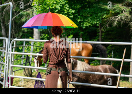 Une jeune femme de race blanche avec des cheveux rouges et vêtements traditionnels est vu tenant un parapluie coloré et debout devant des chevaux Banque D'Images