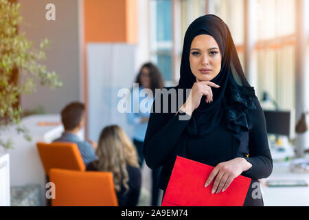 Cheerful woman dans l'office, du dossier avec les documents et de donner un sourire lumineux. L'emploi des femmes musulmanes. Banque D'Images
