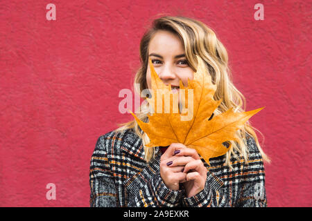 Belle caucasian girl avec un coin dans la feuille d'automne jaune dans les mains sur un arrière-plan d'un mur lumineux. Banque D'Images