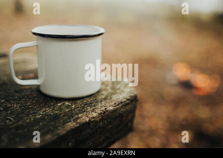 Métal blanc vide mug sur le bord d'une table avec des flous d'un feu de camp à l'arrière-plan Banque D'Images