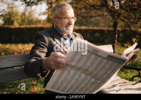 Beau grand-père avec une belle barbe dans un manteau gris s'assied sur un banc dans le parc et lit un journal. Hauts homme aux cheveux gris dans les verres Banque D'Images