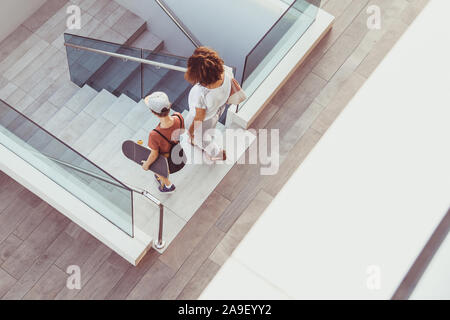 Mère et fils branchés sur des escaliers à pied dans un contexte de centre-ville moderne. Jeune garçon avec skateboard descend l'échelle avec maman vue supérieure de personnes marche dans Banque D'Images