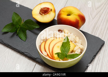 Bol en porcelaine avec le sorgho du porridge, des pêchers, des noix de cajou, d'amandes et de feuilles de menthe sur le conseil de pierre gris et table en bois. Le sorgho sans gluten Vegan Banque D'Images