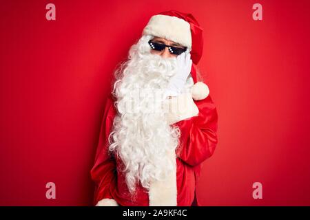 L'âge moyen homme portant un costume de Père Noël et des lunettes de soleil sur fond rouge isolé pensant à fatigué et ennuyé par la dépression Les problèmes avec Banque D'Images