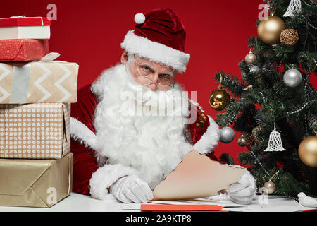 Père Noël assis à la table avec pile de boîtes-cadeaux et la lecture de lettres d'enfants à l'arbre de Noël dans l'arrière-plan Banque D'Images