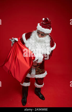 Père Noël en rouge sac et lunettes de holding se présente hors de lui alors qu'il se tenait contre le fond rouge Banque D'Images