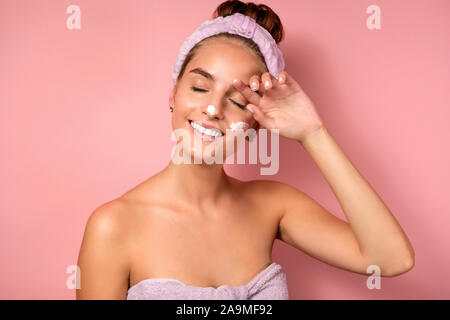 Une fille avec un bandage sur la tête et des correctifs se dresse sur un fond rose avec une crème blanche sur le visage, fermant les yeux et souriant Banque D'Images