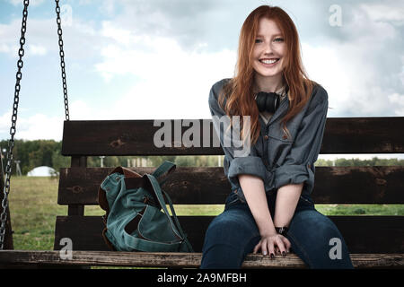 Redhead girl dans une chemise en jean est assis avec un sac à dos sur un banc de bois swing sourit largement à la recherche dans le cadre. Banque D'Images