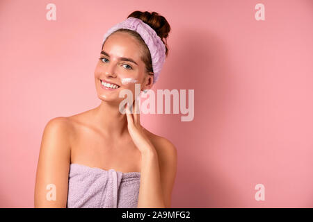 Une fille avec un bandage sur la tête et des correctifs sous ses yeux se dresse sur un fond rose avec une crème blanche sur la joue et sourit Banque D'Images