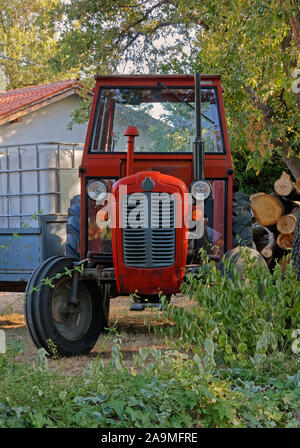 Vieux tracteur rouge avec remorque dans un jardin à côté d'un tas de bois Banque D'Images