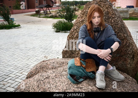 Fille aux cheveux roux est assis sur une pierre avec un sac à dos, jambes croisées et serrant ses genoux avec ses mains, sur une trame de fond du parc. Banque D'Images