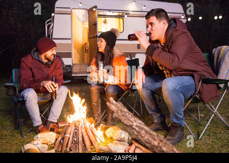 Cheerful friends rire et boire de la bière dans un camp site autour de feu de camp. Retro camping-van. Banque D'Images