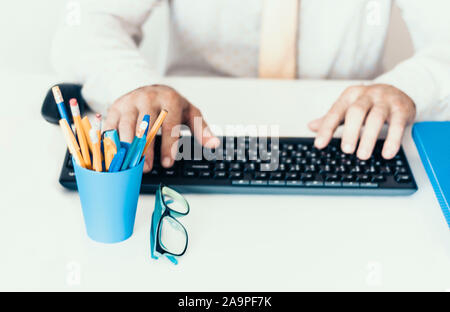 Close-up de mains homme d'âge moyen en chemise blanche en train de taper au clavier de l'ordinateur portable, avec porte-crayons et stylos, pile de livres, cahiers, smartpho Banque D'Images