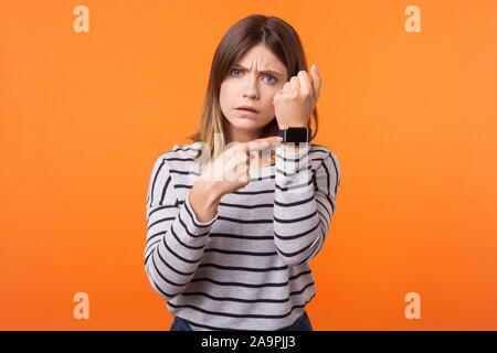 Portrait de jeune femme impatient avec les cheveux bruns en chemise rayée manches longues article pointant sur des montres sur son poignet, à la recherche de sérieux et inquiet. Banque D'Images