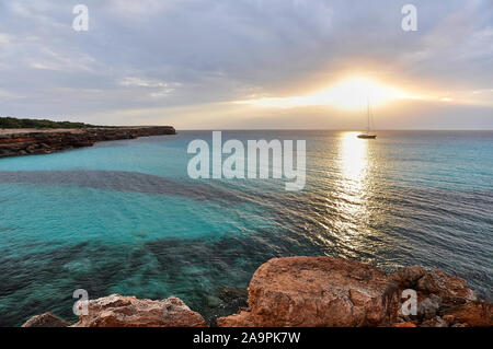 Coucher du soleil à Cala Saona avec soleil rougeoyant et le bleu des eaux cristallines et de voile bateau dans la distance (Îles Baléares, Formentera,Méditerranée,Espagne) Banque D'Images