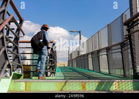 Un voyageur se tient sur un pont et se penche sur la distance. Un homme dans un chapeau et un jean qui voyage avec un sac à dos. Un seul étranger, loin de chez vous. Banque D'Images