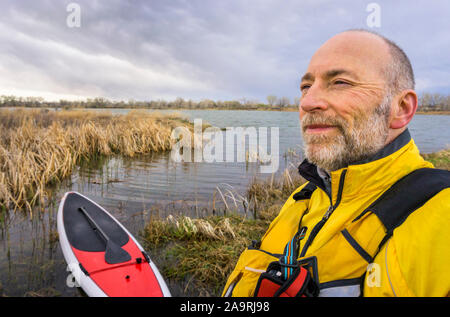 Portrait de l'environnement d'un confiant et heureux pagayeur senior en gilet avec son paddleboard et le lac en arrière-plan, le début du printemps paysage avec Banque D'Images