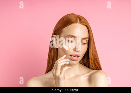 Close up portrait of young woman applying foundation européenne ou d'une crème hydratante sur son visage. Modèle de beauté parfaite avec la peau douce, les taches de rousseur, cheveux roux sur Banque D'Images