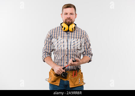 Heureux jeune réparateur barbu de protection avec écouteurs, marteau et ceinture d'outils Banque D'Images