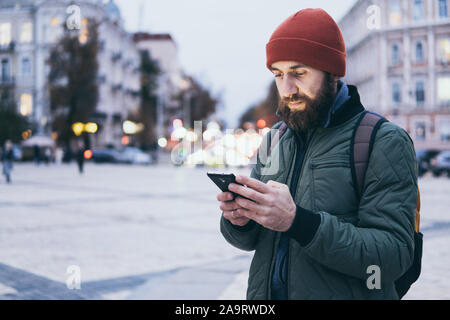 Les jeunes phoques barbus Caucasian man à la recherche sur l'écran du téléphone mobile en marchant sur la rue Banque D'Images