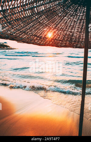 Parasol sur une plage de sable. Soleil à travers l'Osier parapluie à Lever coucher d'été. Banque D'Images