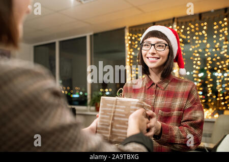 Pretty young woman in Santa cap cértificat de passage à collègue at party Banque D'Images