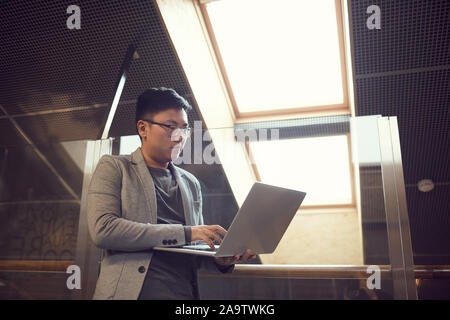 Low angle portrait d'Asian businessman using laptop tout en se tenant dans l'intérieur de bureau moderne, copy space Banque D'Images