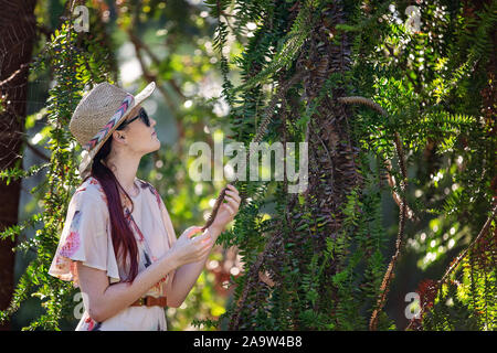 Jolie jeune femme aux longs cheveux rouges portant chapeau de paille, lunettes de soleil et de l'examen d'une robe à fleurs feuilles piquantes Banque D'Images