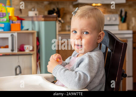 Très jolie petite fille aux yeux bleus, grand, s'asseoir dans sa chaise d'avoir une collation. Banque D'Images