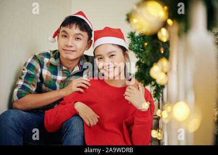 Portrait of happy hugging Asian teenage frère et sœur à Santa hats sitting on steps and looking at camera Banque D'Images