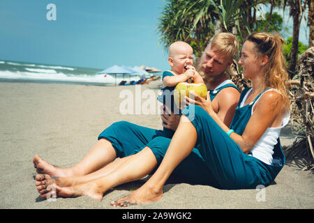 Drôle de famille heureux hipster pique-nique sur plage - mère, père nourrir bébé garçon. Petit-fils de manger des fruits avec plaisir, boire de noix de coco fraîche. Mode de vie sain. Banque D'Images