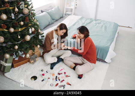 Deux jeunes filles et les ongles des orteils de peinture sur un lit dans le salon près de l'arbre de Noël. Banque D'Images