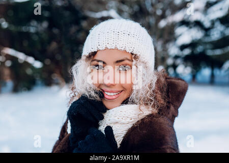 Smiling young woman touching face, portant chapeau d'hiver en tricot blanc élégant et les gants dans winter park. Banque D'Images