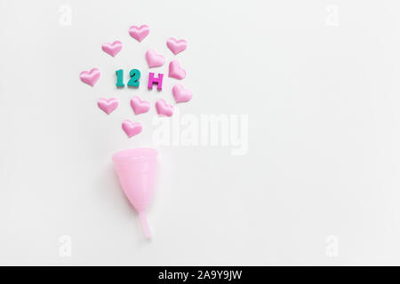 Coupe menstruelle rose sur fond blanc avec des coeurs et la désignation d'utilisation du temps, 12 heures, disposées en nombres en couleur et lettre. Concept zéro déchet, Banque D'Images