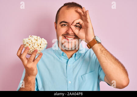 Young man holding bowl with popcorn debout sur fond rose isolé avec happy smiling face ok faire signer avec la main sur les yeux en regardant par la FING Banque D'Images