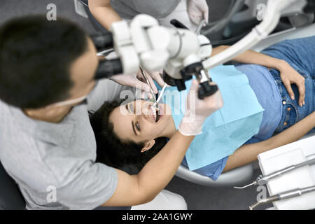 Soins dentiste dans un uniforme gris avec masque de protection et des gants en latex blanc cherche dans un microscope dentaire alors que traite ses female patient in a blue bi Banque D'Images