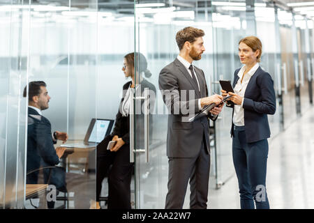 Les gens parler affaires dans le couloir de l'immeuble de bureaux modernes avec les employés qui travaillent derrière le verre des partitions. Travailler dans une grande entreprise Banque D'Images