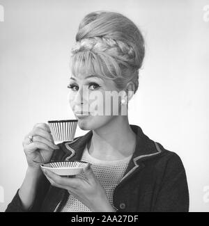 Café dans les années 1960. Une femme est de boire du café à partir d'un 1960 coffeecup avec un motif à rayures. Elle a ses cheveux dans la ruche typique hairstyle, dont les cheveux longs s'entasse sur le dessus de la tête et de donner une certaine ressemblance avec la forme d'une ruche traditionnelle. Suède 1962. Ref BV97-8 Banque D'Images