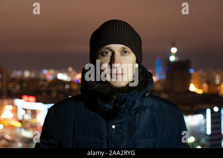 Portrait de la rue d'un jeune homme européen non rasé avec les yeux gris tricoté portant un chapeau chaud et une veste d'hiver chaud, debout dans la nuit sur le toit de Banque D'Images