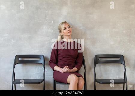 Cute blonde femme en robe rouge est assise sur une chaise et attend que son entrevue. Banque D'Images