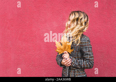 Portrait fille blonde avec les cheveux bouclés est titulaire d'une feuille de coin dans ses mains. L'humeur mélancolique de l'automne. Sur un fond rouge vif. Copier l'espace. Banque D'Images