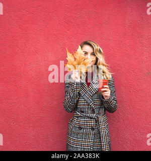 Portrait fille blonde avec les cheveux bouclés est titulaire d'une feuille de coin et une tasse de café pour aller dans ses mains. Sur fond rouge vif. Copier l'espace. Banque D'Images